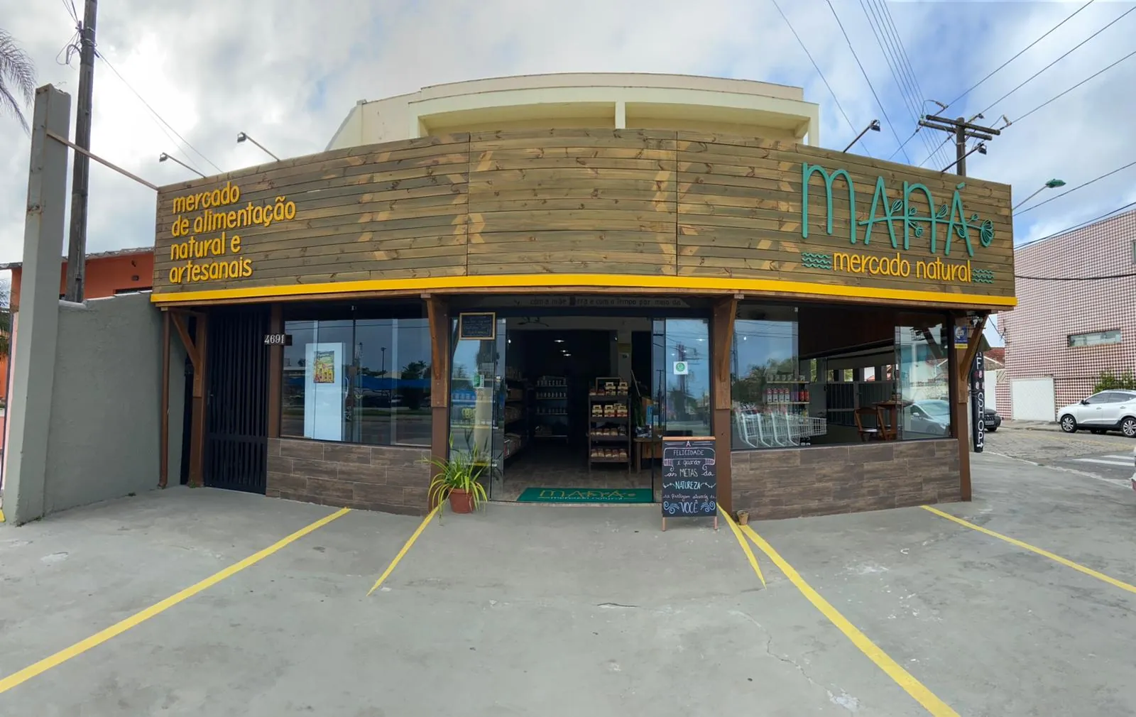 Fachada de madeira do Maná Mercado Natural, com o letreiro verde e amarelo e vagas de estacionamento em frente